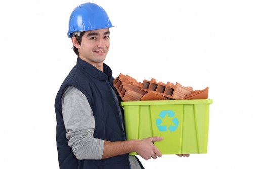 A young man wearing a blue safety helmet and a navy blue vest over a grey long-sleeved shirt is smiling while holding a bright green recycling bin with a blue recycling symbol. The bin contains orange clay bricks stacked neatly, with some portions of the bricks visible at the top. The background is plain white, emphasizing the individual and the waste materials. This scene is relevant to rubbish removal services, illustrating the handling of construction debris or recyclable materials, consistent with services offered by Business Waste Removal Walthamstow. The focus on the person's attire and the clean, organized presentation aligns with professional waste management activities typically carried out in Walthamstow, London, and surrounds. The image communicates a clear message about responsible waste disposal, suitable for a website dedicated to rubbish collection and recycling services in the local area.