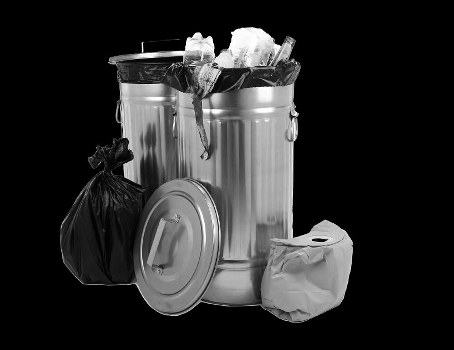 A silver, dome-shaped metal rubbish bin with a lid resting beside it, filled with various waste items including crumpled paper and other household refuse, with a black garbage bag leaning against the bin and a smaller paper bag placed to the right. The scene appears to be set in an indoor or outdoor area near a driveway or pavement, with the waste bin positioned centrally and the background completely black, emphasizing the contents and containers. The overall presentation reflects typical rubbish collection or waste management processes, relevant to services like those offered by Business Waste Removal Walthamstow, operating within the local postcode area and surrounding Walthamstow. The materials, textures, and colors of the waste and containers are clearly depicted to assist visually impaired users and support local SEO signals for rubbish removal in the Walthamstow area.