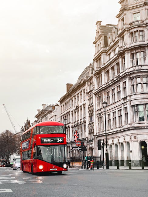 A red double-decker bus traveling along a city street with ornate, multi-story white buildings featuring detailed architectural facades and bay windows in the background. The bus displays route inform