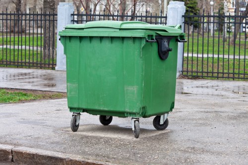 A large green plastic waste bin with a closed lid and four small wheels positioned on a paved surface. The bin is situated outdoors in front of a black metal fence with vertical bars and stone posts, behind which there are trees and a grassy area. The overall scene suggests an urban or suburban environment, possibly near residential or commercial premises in Walthamstow, with natural daylight illuminating the bin and surroundings. The bin appears to be used for rubbish collection and disposal, consistent with services provided by businesses like Business Waste Removal Walthamstow.