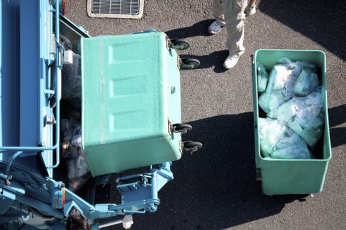 An overhead view of a green waste collection bin filled with clear plastic bags containing various types of rubbish, positioned on a paved surface next to a larger green wheeled refuse container. The image shows part of a blue waste collection vehicle on the left side, with a small section of its metal framework visible. A person’s legs, wearing beige trousers and white shoes, are partially visible at the top of the image, standing near the containers. The environment appears to be outdoors on a paved driveway or street, with the waste containers placed for collection or disposal. The scene emphasizes waste management activities typical of rubbish removal services, such as those provided by Business Waste Removal Walthamstow, and highlights the contents’ translucent plastic bags and the outdoor setting.