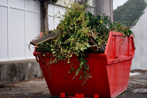 A large, red industrial waste skip filled with mixed green garden waste, including branches, leaves, and clippings, situated outdoors on a paved surface next to a white wall with vertical panels. The waste consists of overgrown shrubbery and foliage, spilling over the edges of the skip, with some small wooden slats and debris mixed in. The background shows a concrete wall, a metal fence or barrier, and a glimpse of trees or shrubbery beyond. The scene is illuminated by natural daylight, emphasizing the texture of the lush green plant matter against the smooth, painted metal surface of the skip, in an environment typical of rubbish removal services supporting garden clearance in Walthamstow or nearby areas. Business Waste Removal Walthamstow regularly handles such garden waste collection, ensuring efficient disposal and management of waste materials like these.