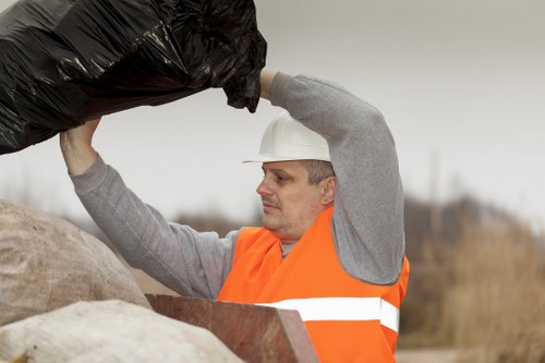 A male waste management worker wearing a white safety helmet and an orange high-visibility vest is lifting a black plastic rubbish bag above his head, preparing to dispose of or transport it. He is standing outdoors in a rural or suburban setting with an overcast sky and blurred natural background, possibly fields or open land. The worker appears focused and is using both hands to hold the rubbish bag, which is filled and tied at the top. The presence of a large pile of mixed rubbish or waste materials, including different bag colors and textures, suggests a collection or removal process typical of waste management services. The image reflects professional rubbish collection activity and visually supports waste disposal operations associated with businesses or community cleanup efforts in the Walthamstow area, managed by companies like Business Waste Removal Walthamstow.